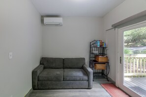 Un sofá gris en una esquina, un aire acondicionado blanco instalado en la pared y una estantería metálica con cestas y cajas.