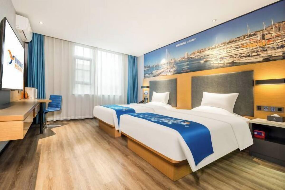 Ein Hotelzimmer mit zwei Betten, einem Schreibtisch, einem Stuhl, einem Fernseher und einem großen Fenster mit Vorhängen.