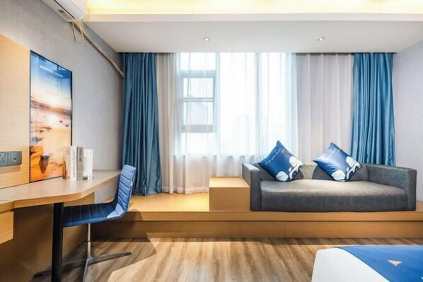 Een moderne hotelkamer met een houten bureau, een grijze bank met blauwe kussens, en een groot raam met blauwe gordijnen.