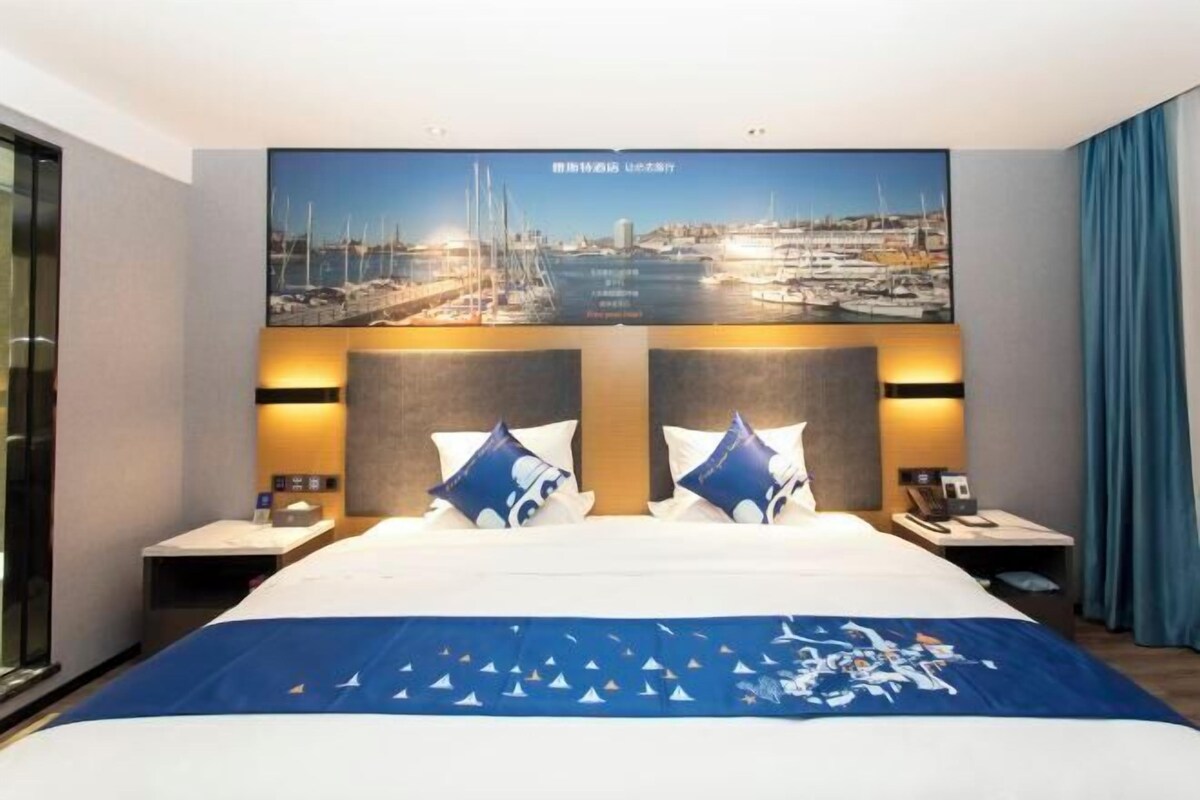A hotel room with a large bed, two nightstands, a flat-screen TV mounted above the bed displaying a marina scene, and a blue curtain on the right.
