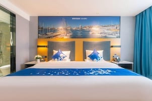 Una habitación de hotel con una cama grande, dos mesitas de noche, un televisor montado en la pared y un cuadro con una vista urbana sobre la cama.