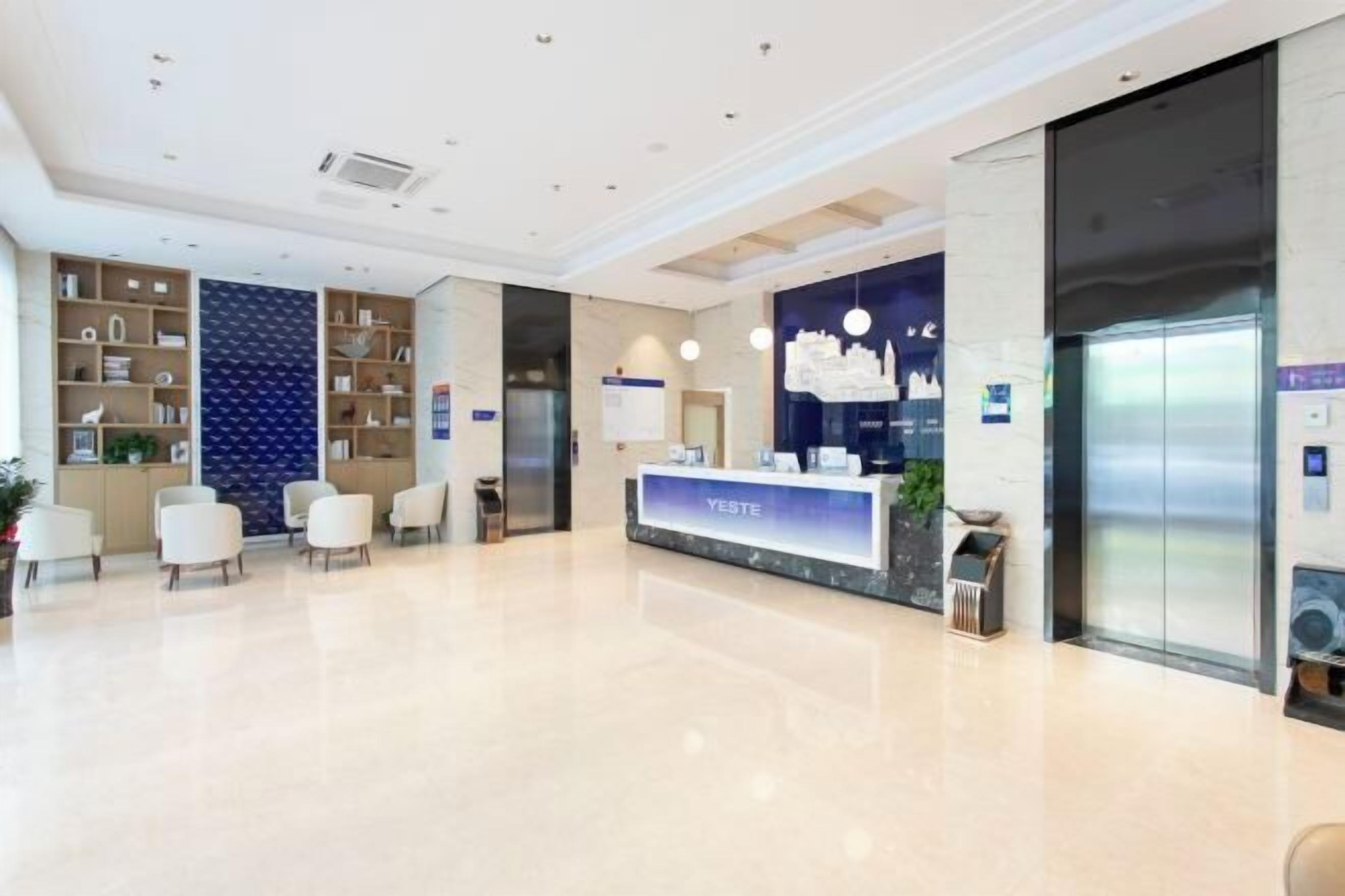 A modern hotel lobby with a reception desk labeled "YESTE," a reception area, seating, and an elevator.