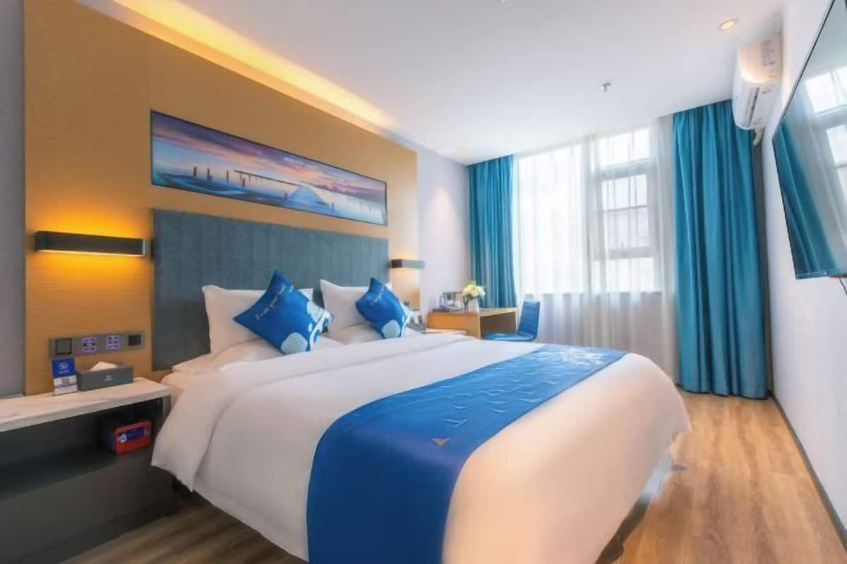 Ein modernes Hotelzimmer mit einem großen Bett, einem Nachttisch mit Lampe, einem Bild an der Wand und einem Fenster mit Vorhängen.