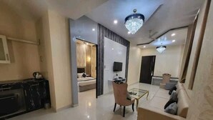 A modern living room with a sofa, a coffee table, and a TV. There is a kitchen area with a black countertop and cabinets. - Corbett View Banquets and Resorts (Moradabad)