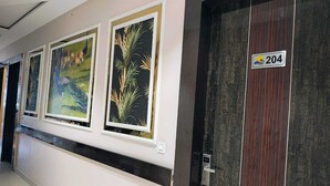 A hallway with framed botanical artwork on the wall, a door with a number sign, and a modern elevator. - Corbett View Banquets and Resorts (Moradabad)