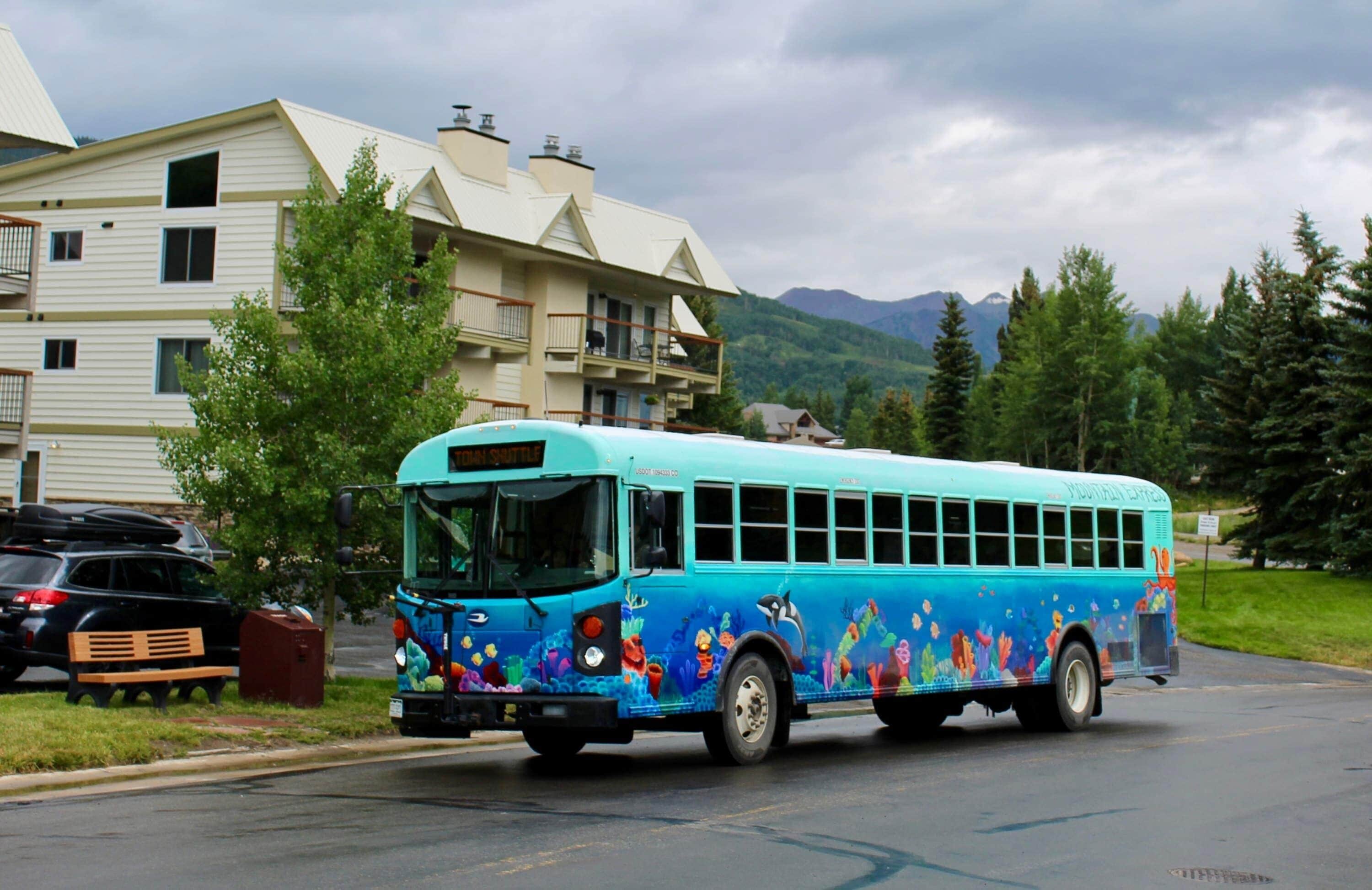 Ein bunter Bus mit maritimem Design, geparkt auf einer Straße, im Hintergrund ein Wohngebäude und ein Berg.