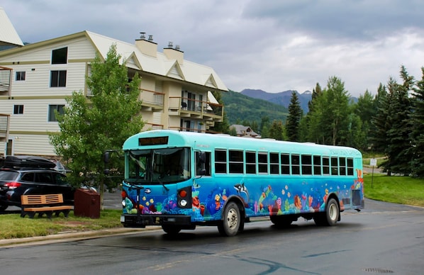 Um ônibus colorido com tema náutico estacionado em uma rua, com um prédio residencial e uma montanha ao fundo.