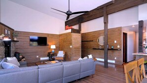 Un saló modern amb un sofà gris, parets de fusta, un televisor de pantalla plana, un ventilador de sostre i una zona de menjador amb cadires de fusta.