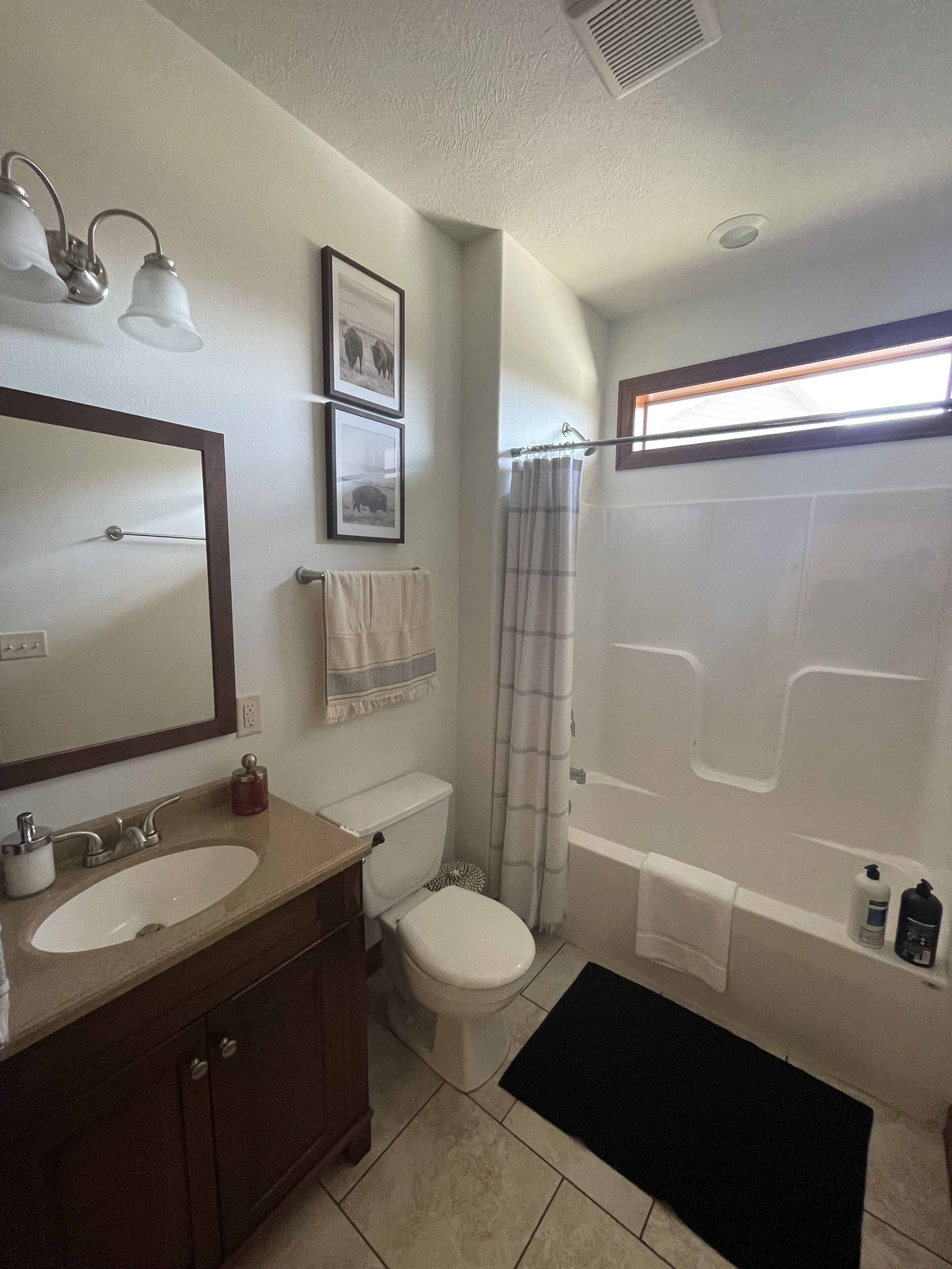 Une salle de bain équipée d’un lavabo, de toilettes et d’une baignoire. Deux appliques murales sont fixées au-dessus du lavabo, une serviette est accrochée au mur et un rideau de douche est installé.