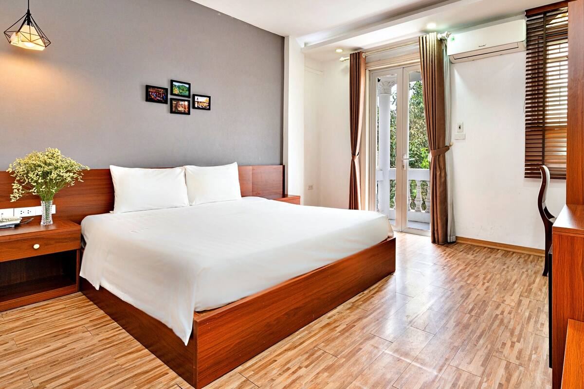 A hotel room with a large bed, wooden flooring, a nightstand with a vase of flowers, a desk with a fruit tray, and a wall with framed pictures.