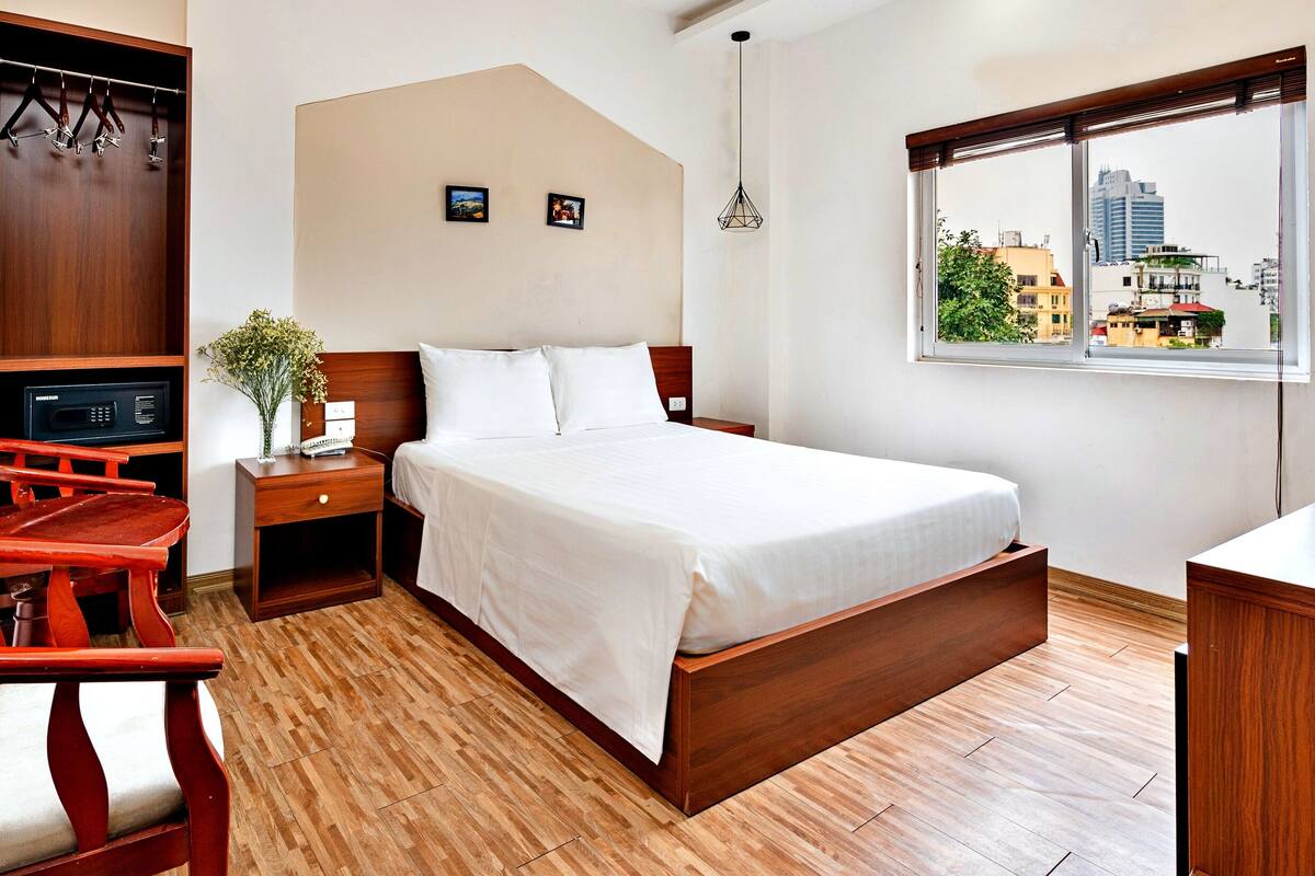 A hotel room with a bed, a wooden nightstand, a chair, a desk, a window with a city view, and a wall-mounted TV.