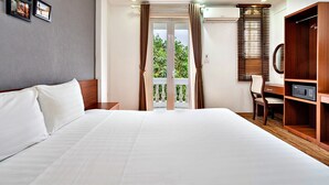Une chambre d’hôtel avec un grand lit, une armoire en bois, un bureau avec une chaise et une vue sur la verdure par les portes du balcon.