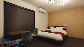 A hotel room with a bed, a desk, a chair, and an air conditioning unit.