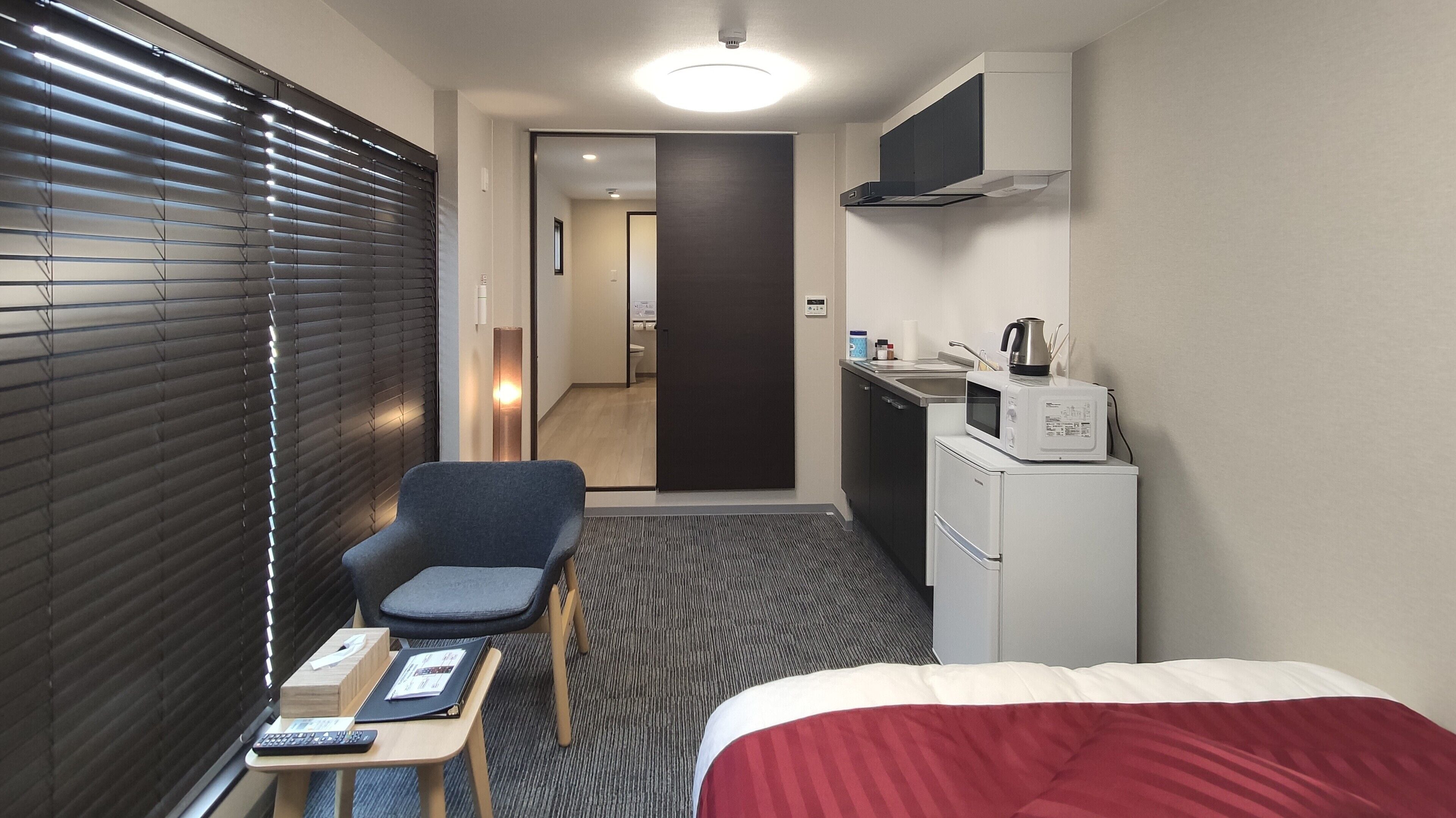 A hotel room with a bed, a desk with a chair, a kitchenette with a microwave, and a door leading to a bathroom.