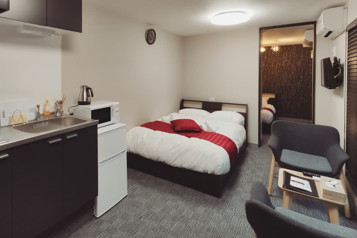 A compact hotel room with a kitchenette, a bed with red and white bedding, a small desk, and a chair.