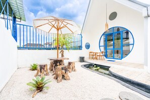 Ein Innenhof mit Holztisch und -stühlen, einem kleinen Wasserspiel und einem blau gerahmten Fenster.