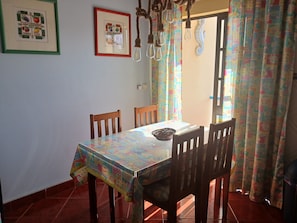 Un coin repas avec une table recouverte d’une nappe colorée, des chaises en bois, un lustre et un rideau à motifs.