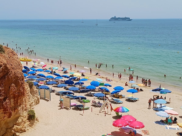 Une plage parsemée de nombreux parasols et de chaises longues, un grand navire de croisière en arrière-plan et une falaise rocheuse sur la gauche.