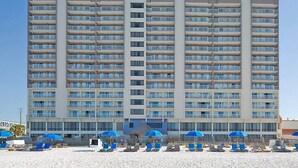 A multi-story hotel with balconies, a beach area with sun loungers and umbrellas, and a clear blue sky.