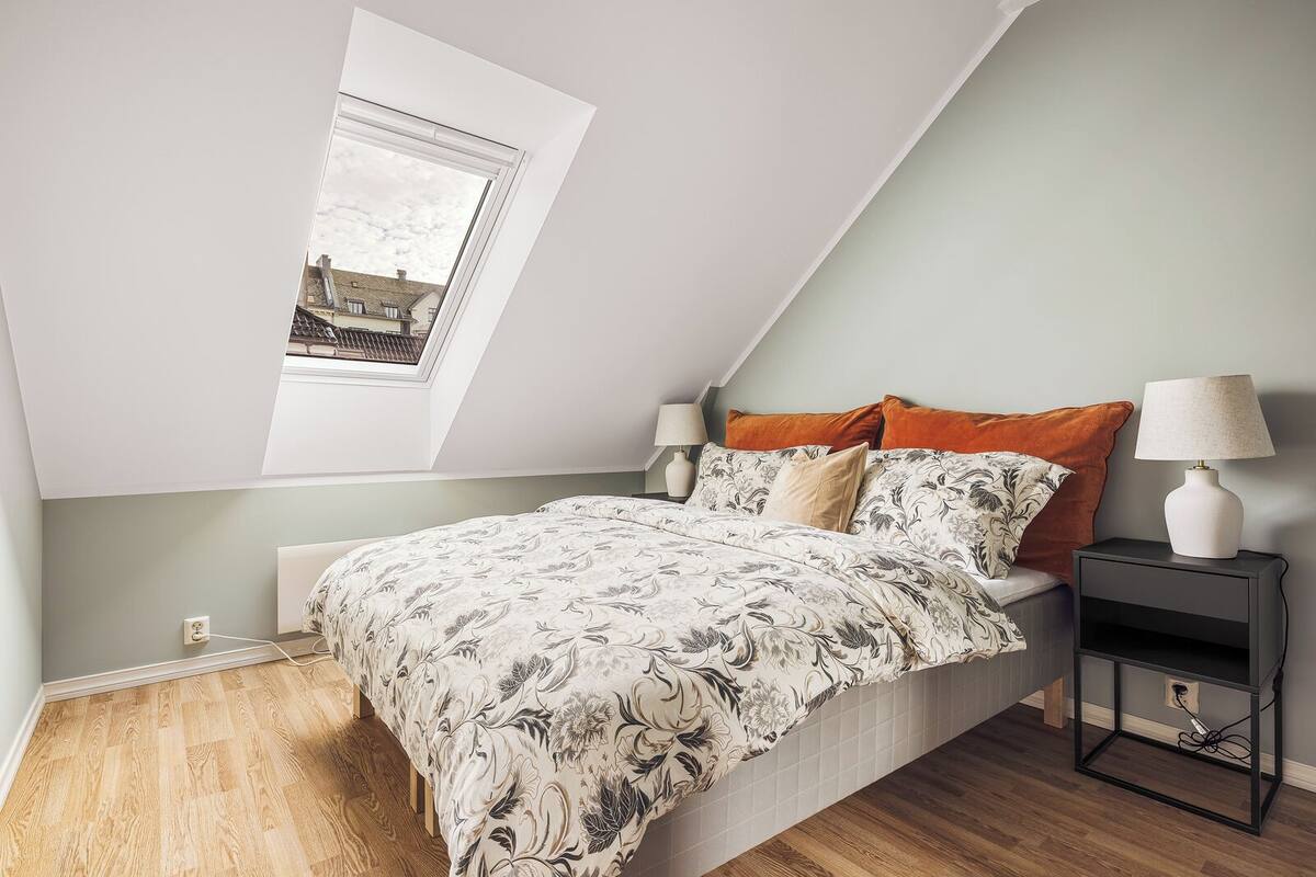 A bedroom with a sloped ceiling, a skylight, a bed with floral bedding, two bedside lamps, and a nightstand with a lamp.