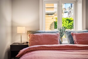 Un dormitorio con cama, almohadas, una mesita de noche, lámpara y ventana con vistas a edificios y follaje.