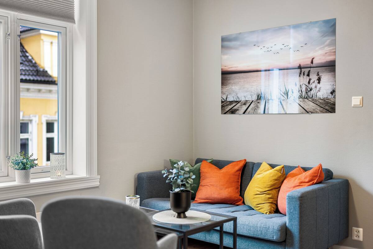 A modern living room with a grey sofa, colorful cushions, a small table with a plant, and a large painting of a sunset over water.