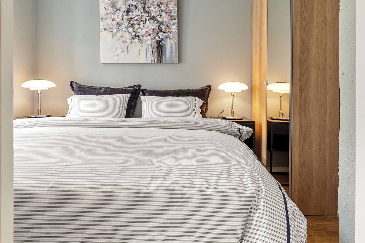 A neatly made bed with striped bedding, two bedside lamps, and a large abstract painting on the wall.