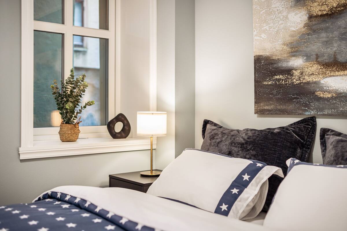 A neatly made bed with a star-patterned blanket, a bedside lamp, a potted plant, and a wall art piece.