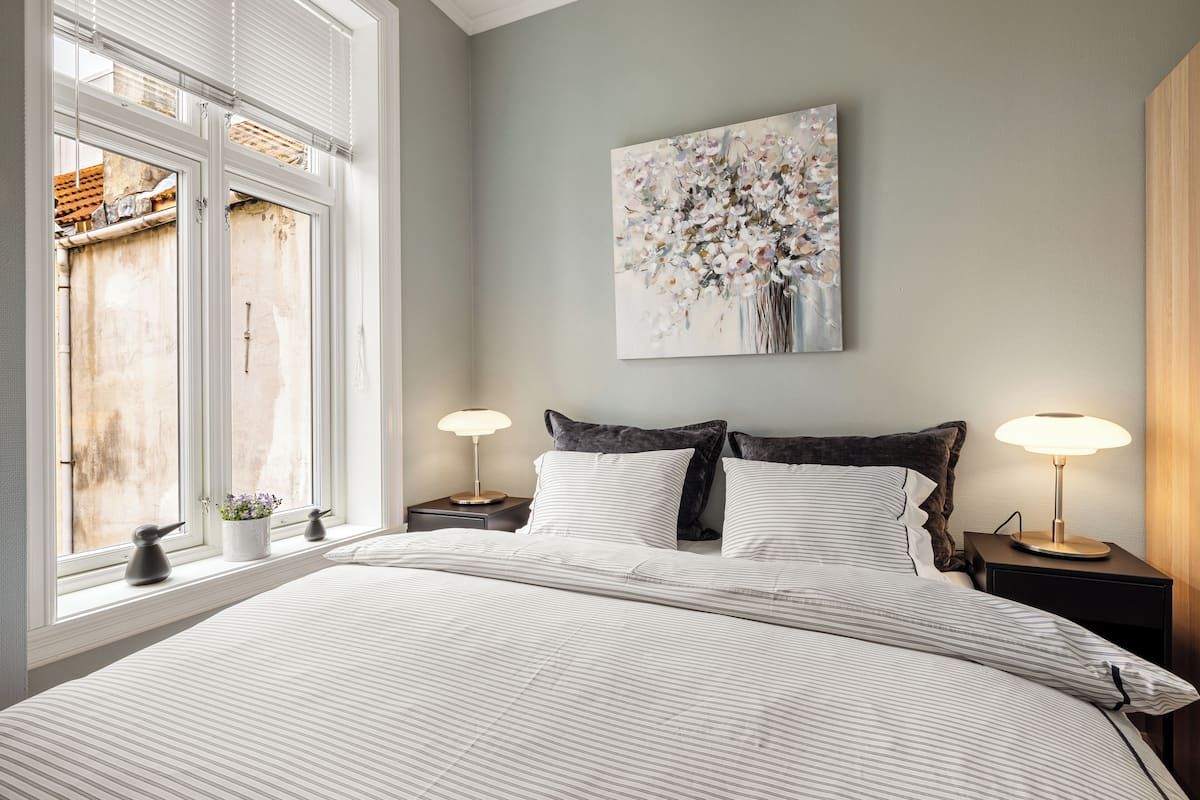 A bedroom with a large bed, two bedside tables with lamps, a window with blinds, and a wall-mounted painting.