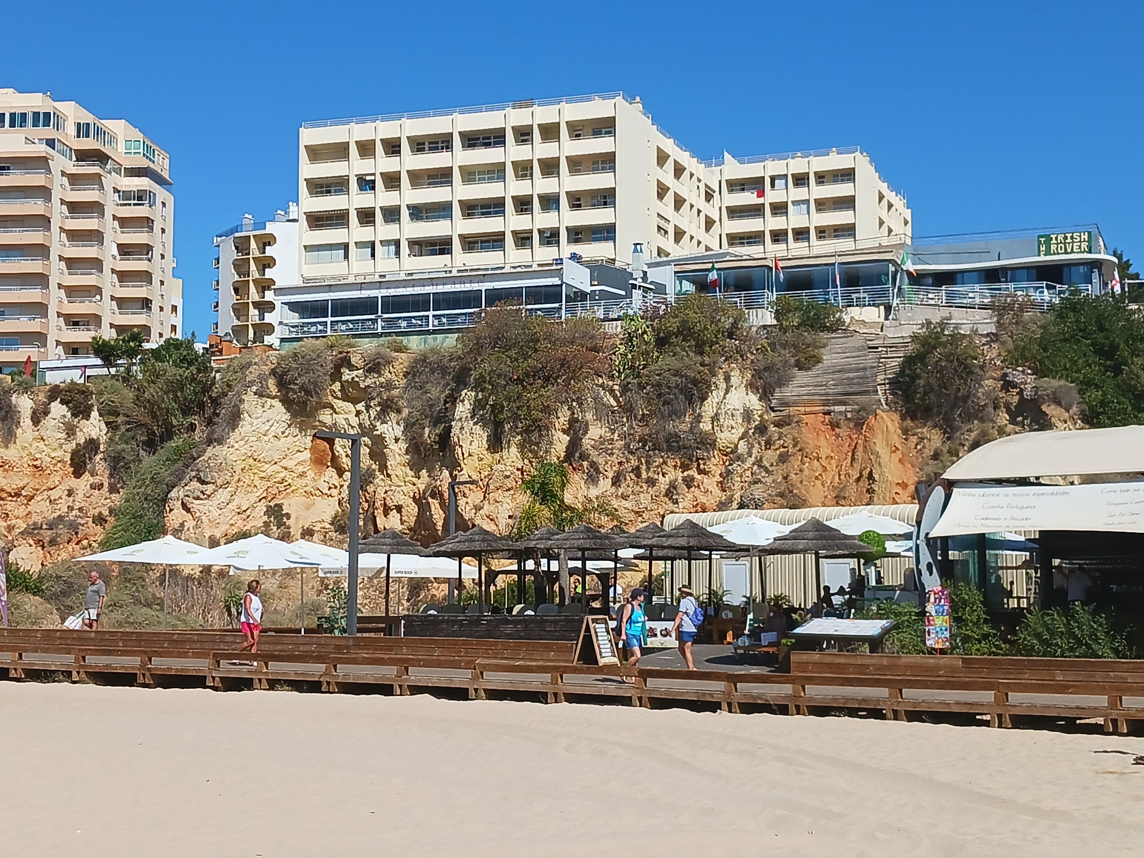 A coastal area with a rocky cliff, a beach, and a row of buildings including a hotel with a sign reading "IRISH ROVER".