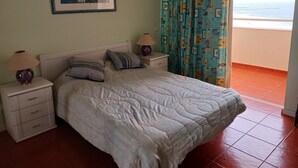 Een slaapkamer met een bed, twee nachtkastjes, een lamp, een ingelijste foto aan de muur en uitzicht op de oceaan door een raam.