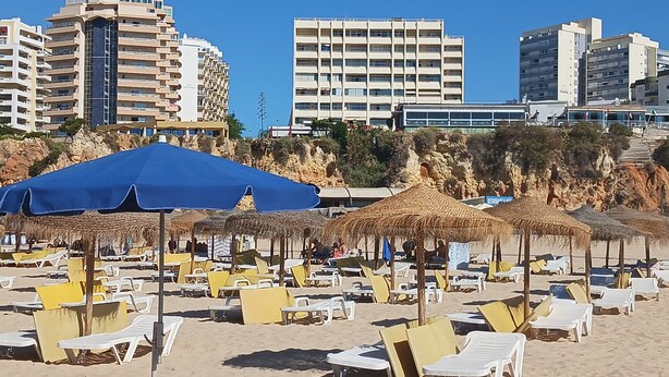 Een strand met rijen ligbedden en rieten parasols, een rotsklif en moderne gebouwen op de achtergrond.