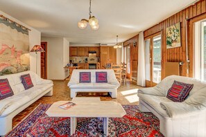 Uma sala de estar ampla com dois sofás, uma mesa de centro e um tapete grande. Dá para ver uma cozinha ao fundo, com armários de madeira e um espaço para refeições.