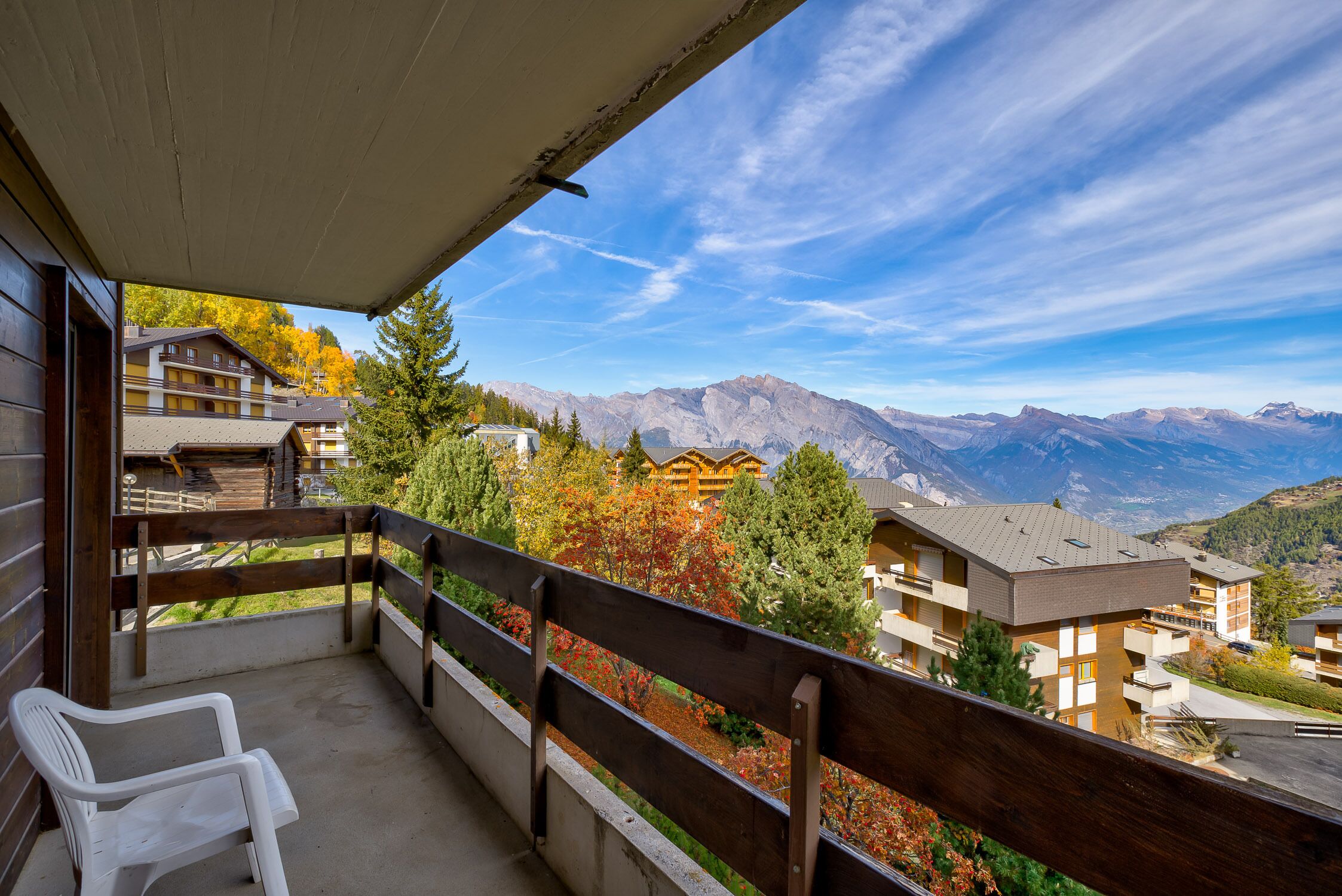 A balcony with a wooden railing, a white plastic chair, and a view of mountain buildings and trees.