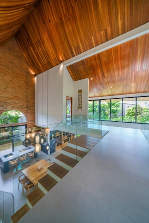 A spacious interior with a high ceiling, modern furnishings, and large windows offering a view of greenery outside. - Villa Satiya (Munggu)