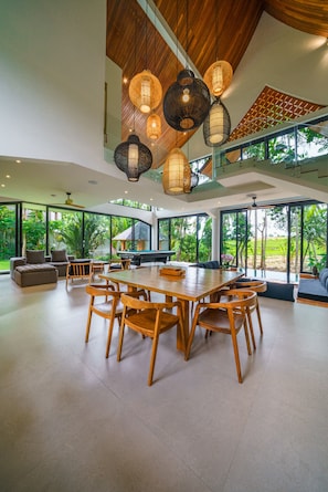 A spacious living area with a large wooden dining table surrounded by chairs, a sofa, and a pool table. The room features large windows offering views of greenery outside. - Villa Satiya (Munggu)