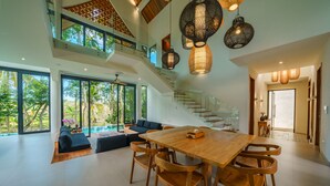A spacious living area with a large wooden dining table and chairs, a staircase, and large windows offering a view of the outdoors. - Villa Satiya (Munggu)
