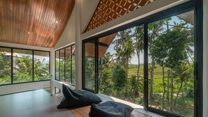 A spacious room with large windows offering a view of lush greenery and palm trees. - Villa Satiya (Munggu)