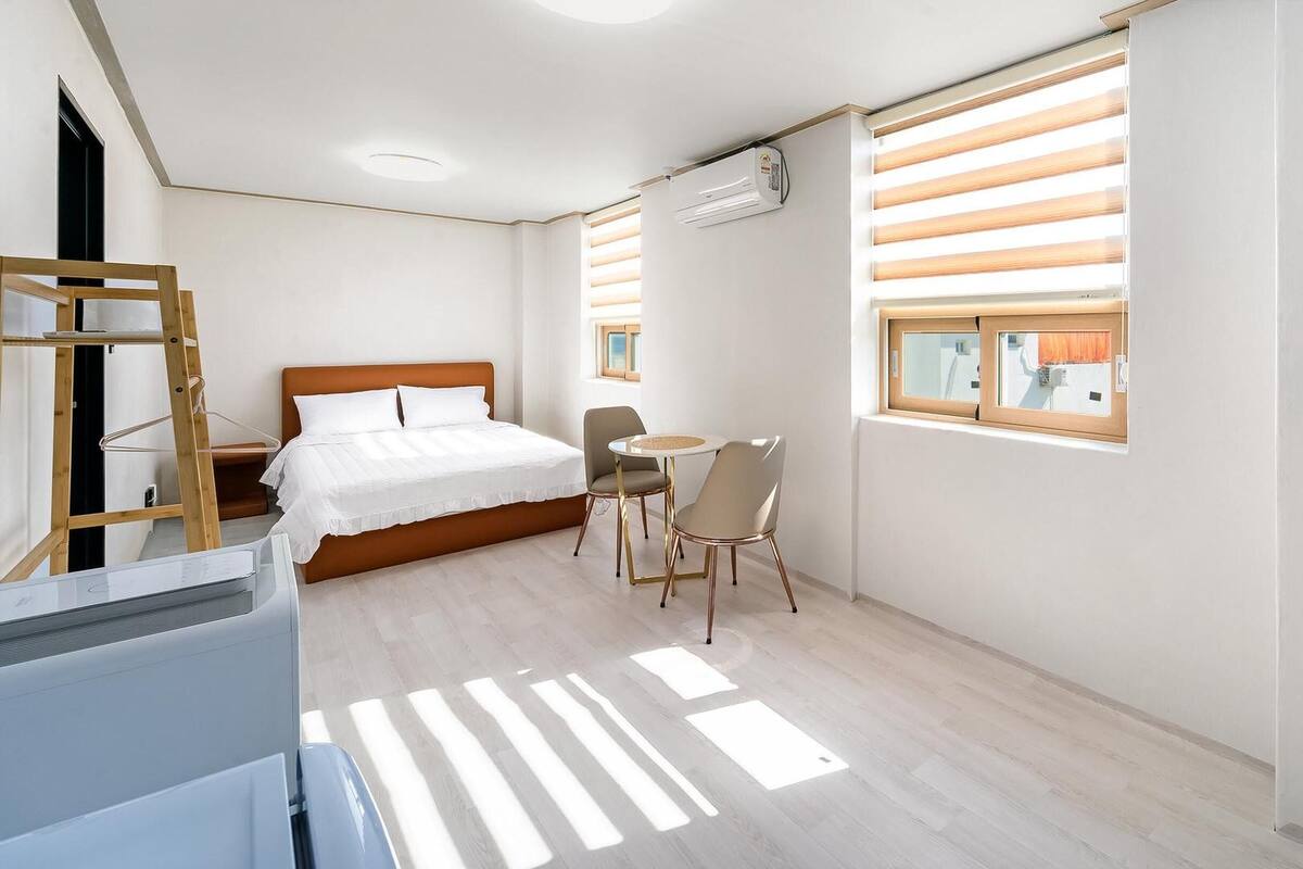 Un dormitorio con cama, escritorio y silla. Hay una ventana con persianas, un equipo de aire acondicionado y un televisor montado en la pared.
