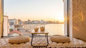 Ein Zimmer mit Blick auf Strand und Stadtlandschaft, ein Tisch mit Teekanne und Tassen sowie zwei gewebte Poufs auf einer gepolsterten Bank.