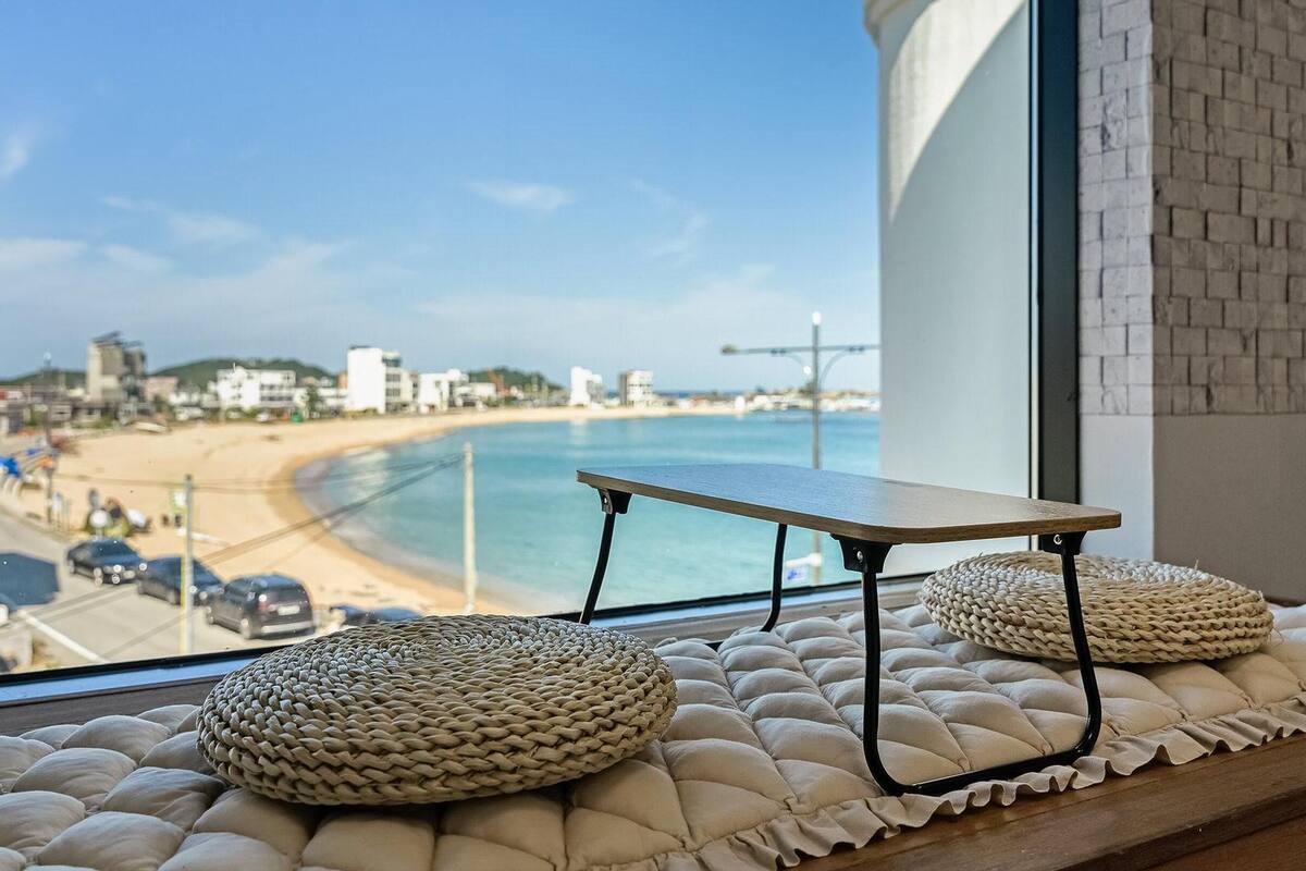 Vista de una playa desde una ventana, con una mesa y dos otomanas tejidas.