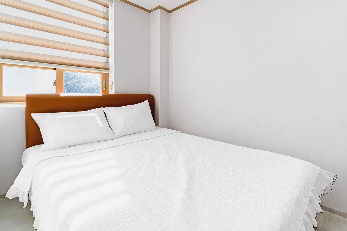 Una cama bien hecha con ropa blanca en una habitación con cabecera de madera y una ventana con persianas.