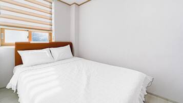 A neatly made bed with white bedding in a room with a wooden headboard and a window with blinds.