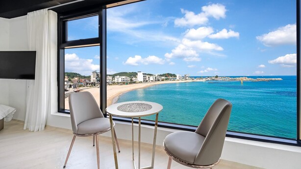 Habitació amb finestra gran que dona a una platja i a l'oceà, dues cadires i una taula petita.