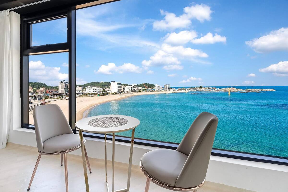 Una habitación con un amplio ventanal que da a una playa y al océano, dos sillas y una mesita.