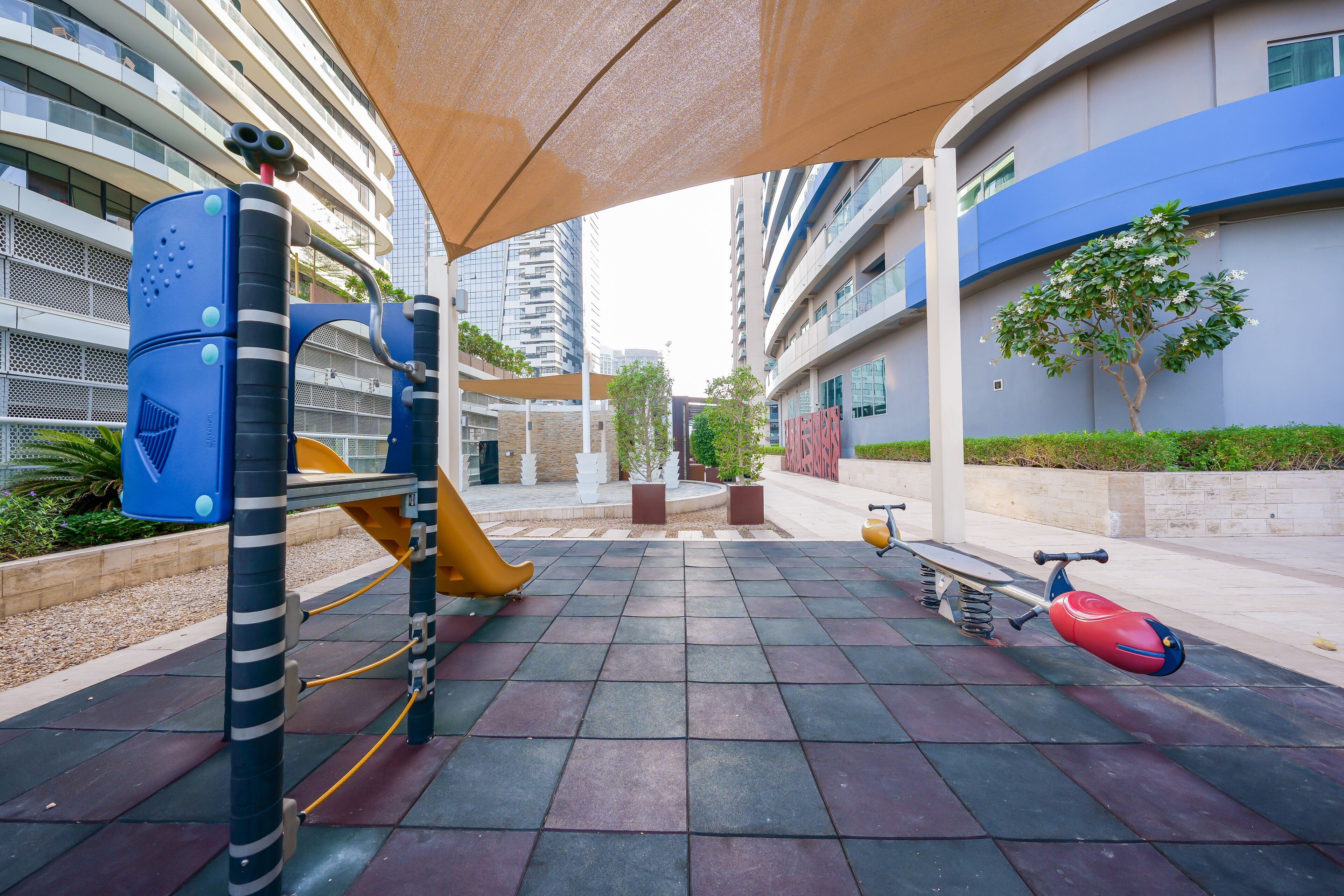 Un terrain de jeux avec un toboggan et des balançoires, entouré de bâtiments modernes et d’un arbre.