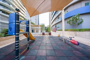 Un terrain de jeux avec un toboggan et des balançoires, entouré de bâtiments modernes et d’un arbre.