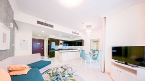 Una sala de estar moderna con un sofá, una mesa de centro y un televisor. Se ve un área de cocina al fondo, con una mesa de comedor y sillas.