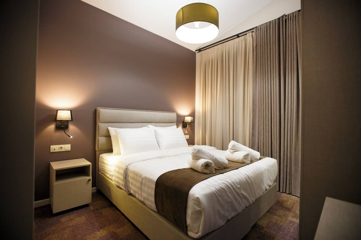 A hotel room with a bed, nightstands, wall-mounted lamps, and a window with curtains.