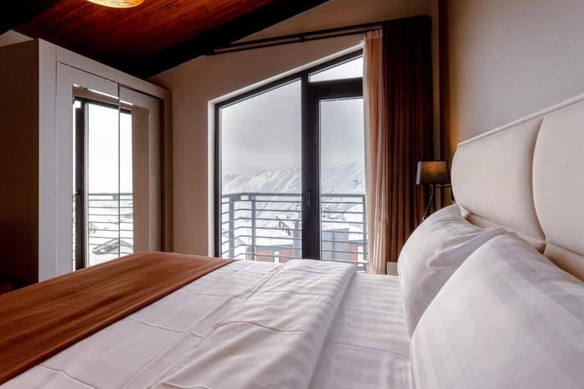 A hotel room with a bed, a view of mountains, and a balcony with a railing.